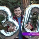 Smiling man holding large silver number balloons "3" and "0" outdoors.